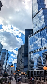 Tall skyscrapers dominate the cityscape under a partly cloudy sky. The glass buildings reflect the surrounding structures and the sky. A prominent building with the text 'TRUMP' is visible in the foreground. Street lights with orange globes line the walkway where people are walking.