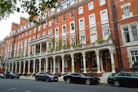 A row of elegant, historic brick buildings featuring ornate architectural details, large windows, and a covered balcony with pillars. The street is lined with several parked cars and has lush greenery on the building's balcony and nearby trees.