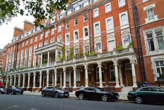 A row of elegant, historic brick buildings featuring ornate architectural details, large windows, and a covered balcony with pillars. The street is lined with several parked cars and has lush greenery on the building's balcony and nearby trees.