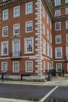 A multi-story red brick building with white-framed windows. The structure has decorative stonework along the edges and a small balcony on the second floor. A street sign indicates the name is 'Grosvenor Street.' A person wearing a blue jacket is walking on the sidewalk parallel to the building. There is a black metal fence bordering the property, small paved courtyard, and a well-maintained sidewalk.