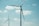 Two large wind turbines stand against a bright sky with scattered clouds. The blades of the turbines are clearly visible, adding to the sense of scale and height. The scene suggests a focus on renewable energy.