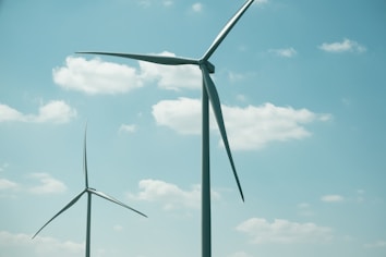 Two large wind turbines stand against a bright sky with scattered clouds. The blades of the turbines are clearly visible, adding to the sense of scale and height. The scene suggests a focus on renewable energy.