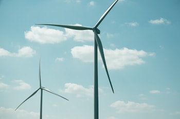 Two large wind turbines stand against a bright sky with scattered clouds. The blades of the turbines are clearly visible, adding to the sense of scale and height. The scene suggests a focus on renewable energy.