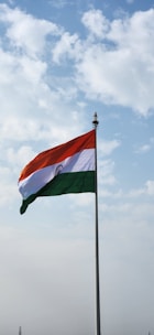 An Indian flag waves proudly against a backdrop of a partly cloudy sky. The flag features horizontal stripes of saffron, white, and green, with a blue Ashoka Chakra in the center of the white stripe.
