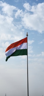 An Indian flag waves proudly against a backdrop of a partly cloudy sky. The flag features horizontal stripes of saffron, white, and green, with a blue Ashoka Chakra in the center of the white stripe.