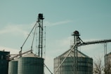 Two large industrial silos with metal structures and conveyor belts against a clear sky. The machinery and support beams suggest an agricultural or industrial setting.