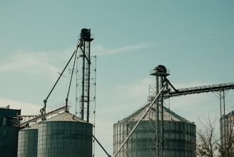 Two large industrial silos with metal structures and conveyor belts against a clear sky. The machinery and support beams suggest an agricultural or industrial setting.