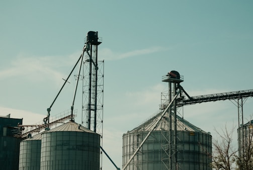 Two large industrial silos with metal structures and conveyor belts against a clear sky. The machinery and support beams suggest an agricultural or industrial setting.