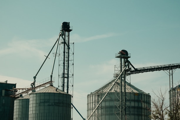 Two large industrial silos with metal structures and conveyor belts against a clear sky. The machinery and support beams suggest an agricultural or industrial setting.