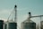 Two large industrial silos with metal structures and conveyor belts against a clear sky. The machinery and support beams suggest an agricultural or industrial setting.