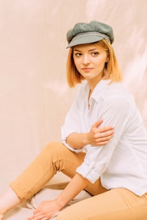 A person with short blonde hair is wearing a white shirt and beige pants while sitting in a relaxed pose. They have a calm and neutral expression on their face, and a green hat adds a stylistic touch. The background is soft and slightly textured, contributing to a serene atmosphere.