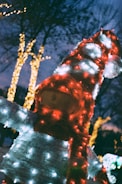 A festive outdoor decoration resembling a snowman-like figure is adorned with numerous colorful holiday lights. The lights illuminate the figure's body with shades of red and white, while strings of yellow lights twinkle on trees in the background under the evening sky.
