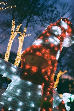 A festive outdoor decoration resembling a snowman-like figure is adorned with numerous colorful holiday lights. The lights illuminate the figure's body with shades of red and white, while strings of yellow lights twinkle on trees in the background under the evening sky.