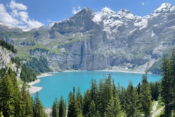 A stunning alpine landscape featuring a turquoise lake surrounded by lush green pine trees. Towering snow-capped mountains provide a majestic backdrop under a clear blue sky with a few scattered clouds.