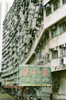 A large, densely packed apartment building is shown with numerous air conditioning units and laundry hanging from the balconies. The structure curves horizontally, adding a dynamic perspective to the scene. Below the building, there is a street with several people walking and a bright green sign featuring large red Chinese characters.