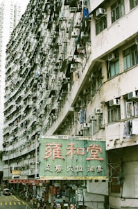 A large, densely packed apartment building is shown with numerous air conditioning units and laundry hanging from the balconies. The structure curves horizontally, adding a dynamic perspective to the scene. Below the building, there is a street with several people walking and a bright green sign featuring large red Chinese characters.