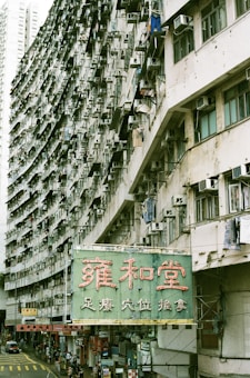 A large, densely packed apartment building is shown with numerous air conditioning units and laundry hanging from the balconies. The structure curves horizontally, adding a dynamic perspective to the scene. Below the building, there is a street with several people walking and a bright green sign featuring large red Chinese characters.