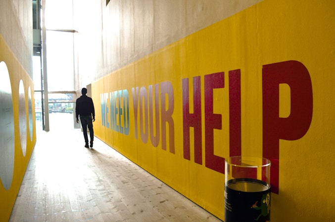 A person walking down a narrow corridor with sunlight streaming in from large windows. The walls are painted yellow with large text that reads 'WE NEED YOUR HELP'. A transparent display case is visible in the foreground.