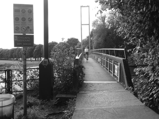 A cyclist rides across a narrow footbridge surrounded by lush trees and vegetation. A sign post with various instructions is visible near the entrance to the bridge. The surroundings include a well-lit pathway and a trash can nearby. The air seems fresh, with trees lining a distant park area.
