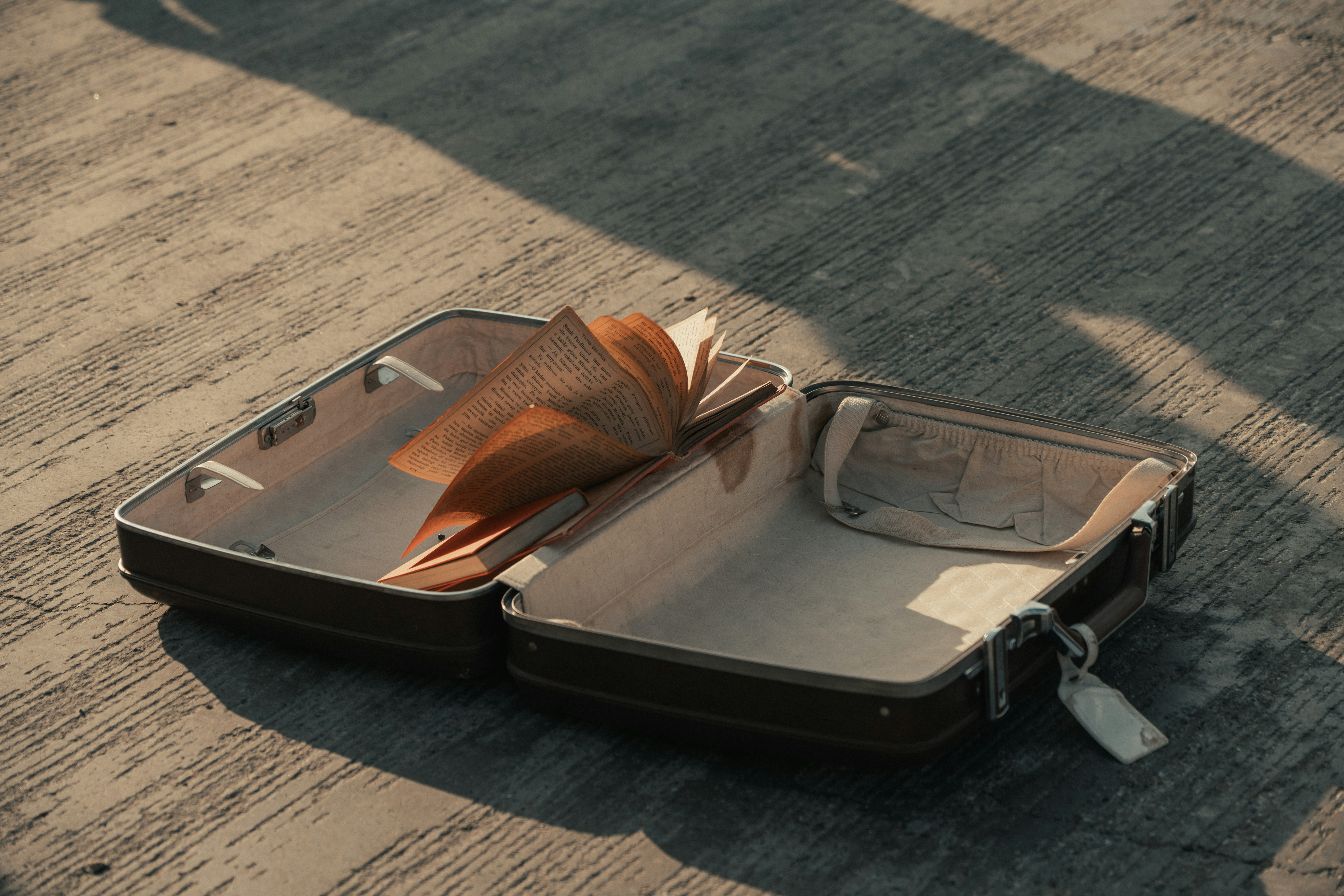 Open vintage suitcase on pavement with a book and travel items in warm sunlight