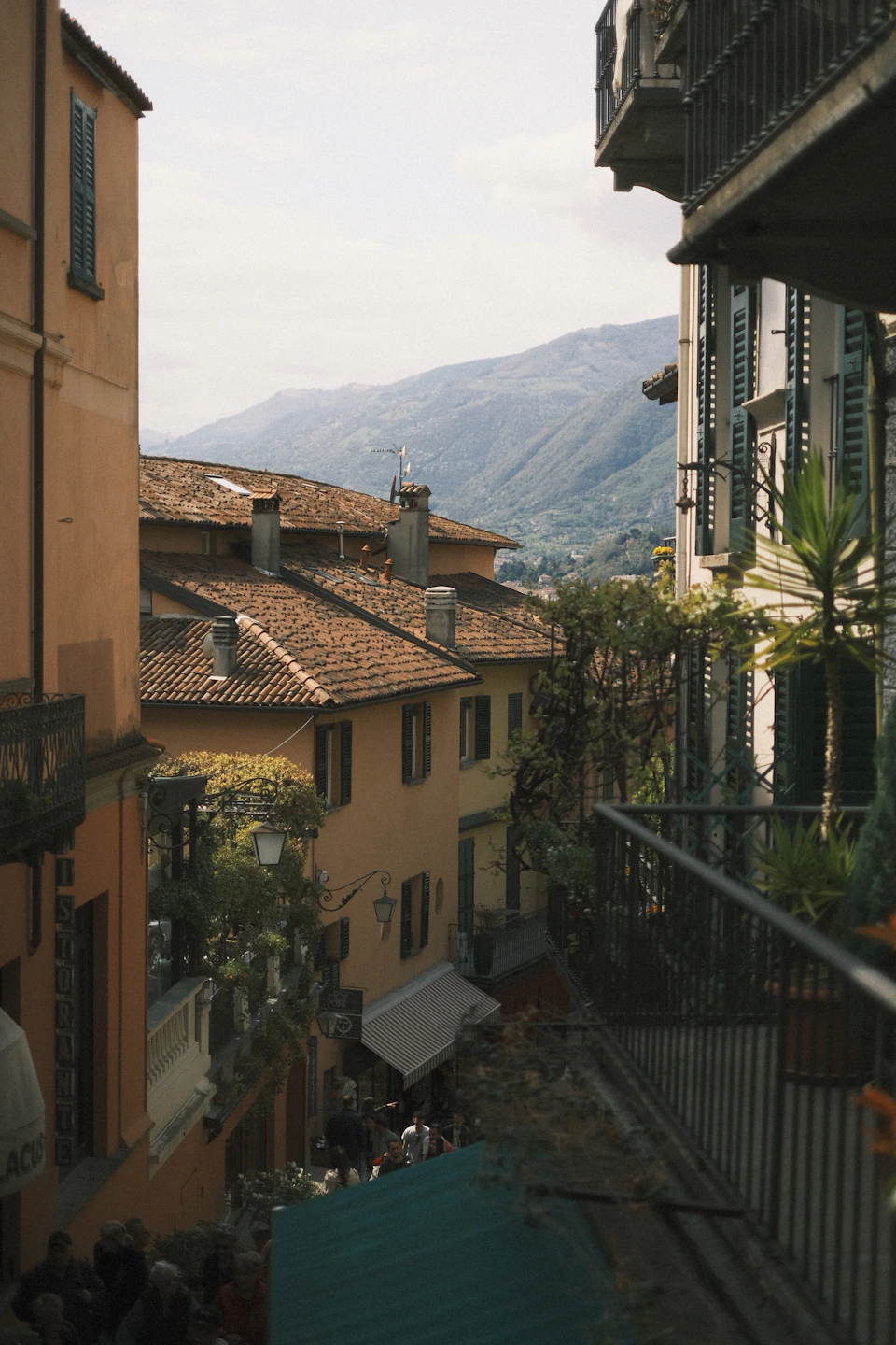 The image depicts a picturesque street scene with buildings on both sides, each boasting unique architectural styles and colors. The buildings are painted in shades of orange and white, creating a warm and inviting atmosphere. On one side of the street, there is a balcony adorned with green plants that adds a touch of nature to the urban setting.

The street itself is bustling with activity, as several people can be seen walking along it or standing on its edge. The perspective of the image offers a panoramic view of the street and the surrounding landscape, which includes mountains in the distance.
