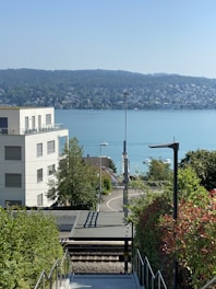 A scenic view of a tranquil lakeside area with a modern apartment building in the foreground. The scene includes a staircase leading down to a railway platform, lush greenery on either side, and a clear blue lake with hills in the background under a bright sky.