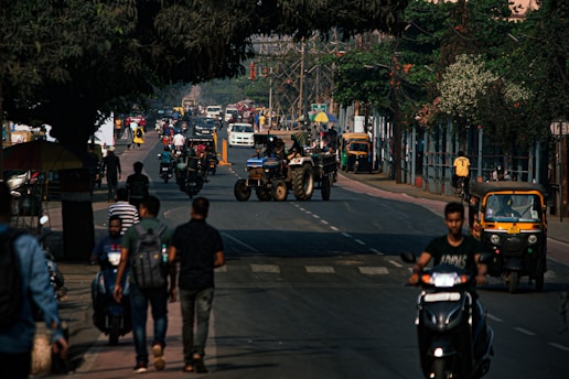 A bustling urban street with a variety of vehicles including motorcycles, an auto rickshaw, and a tractor. There are multiple people walking on the sidewalks and trees lining the street. The atmosphere is busy with a mix of pedestrians and vehicular movement.