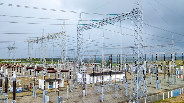 A complex network of electrical infrastructure composed of metal towers, transformers, wires, and high voltage equipment, set against a partially cloudy sky. The landscape includes green vegetation in the background and gravel areas around the structures.
