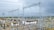 A complex network of electrical infrastructure composed of metal towers, transformers, wires, and high voltage equipment, set against a partially cloudy sky. The landscape includes green vegetation in the background and gravel areas around the structures.