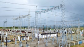 A complex network of electrical infrastructure composed of metal towers, transformers, wires, and high voltage equipment, set against a partially cloudy sky. The landscape includes green vegetation in the background and gravel areas around the structures.