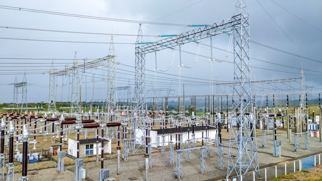 A complex network of electrical infrastructure composed of metal towers, transformers, wires, and high voltage equipment, set against a partially cloudy sky. The landscape includes green vegetation in the background and gravel areas around the structures.