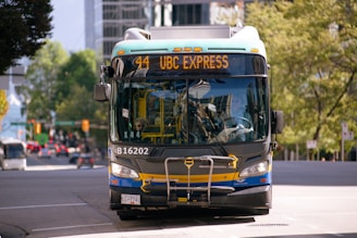 A public transit bus marked as 44 UBC EXPRESS is centered in the frame. The background includes urban elements such as buildings and traffic lights, while trees add a touch of greenery. The bus is painted with blue and yellow accents.