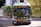 A public transit bus marked as 44 UBC EXPRESS is centered in the frame. The background includes urban elements such as buildings and traffic lights, while trees add a touch of greenery. The bus is painted with blue and yellow accents.
