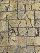 A close-up view of a weathered stone pavement with cracks and uneven surfaces, consisting of rectangular tiles. The stones show signs of aging and erosion, with visible rough textures and varying shades of brown and gray.