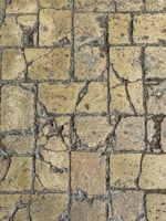 A close-up view of a weathered stone pavement with cracks and uneven surfaces, consisting of rectangular tiles. The stones show signs of aging and erosion, with visible rough textures and varying shades of brown and gray.