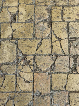 A close-up view of a weathered stone pavement with cracks and uneven surfaces, consisting of rectangular tiles. The stones show signs of aging and erosion, with visible rough textures and varying shades of brown and gray.