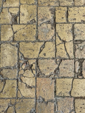 A close-up view of a weathered stone pavement with cracks and uneven surfaces, consisting of rectangular tiles. The stones show signs of aging and erosion, with visible rough textures and varying shades of brown and gray.