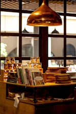 A cozy interior with a large copper pendant light hanging from the ceiling. Below the light, a wooden counter displays neatly stacked cookbooks and cutting boards. In the background, jars filled with various food items are visible. The surrounding atmosphere is warm and inviting, with a focus on culinary themes.