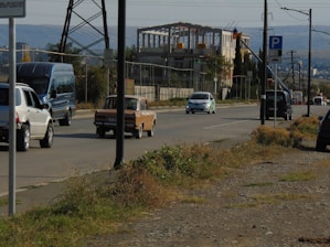 A road scene includes several cars driving along a paved street. A blue van, a white SUV, and an older brown sedan are among the vehicles visible. To the right, a multi-story building under construction is surrounded by safety fencing. The foreground has patches of grass and dry vegetation beside the road. Utility poles and electric wires are along the road, with a parking sign visible.