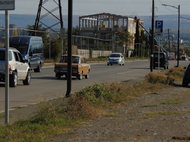 A road scene includes several cars driving along a paved street. A blue van, a white SUV, and an older brown sedan are among the vehicles visible. To the right, a multi-story building under construction is surrounded by safety fencing. The foreground has patches of grass and dry vegetation beside the road. Utility poles and electric wires are along the road, with a parking sign visible.