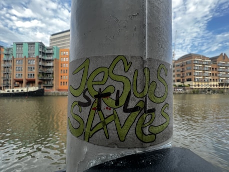 A graffiti sticker with the words 'Jesus Saves' is affixed to a vertical pole. The background features a river with residential buildings and a boat docked alongside. The sky is partly cloudy with hints of blue.