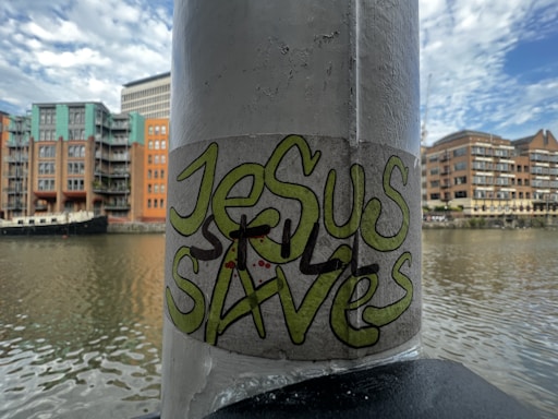A graffiti sticker with the words 'Jesus Saves' is affixed to a vertical pole. The background features a river with residential buildings and a boat docked alongside. The sky is partly cloudy with hints of blue.