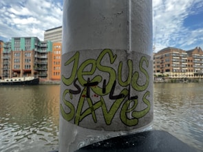 A graffiti sticker with the words 'Jesus Saves' is affixed to a vertical pole. The background features a river with residential buildings and a boat docked alongside. The sky is partly cloudy with hints of blue.