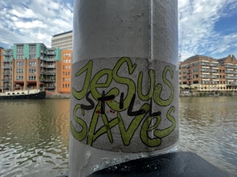 A graffiti sticker with the words 'Jesus Saves' is affixed to a vertical pole. The background features a river with residential buildings and a boat docked alongside. The sky is partly cloudy with hints of blue.
