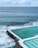 An ocean pool is set next to crashing waves, with several swimmers enjoying the water. The pool is bordered by a white edge and has a natural ocean backdrop with slightly cloudy skies.