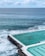 An ocean pool is set next to crashing waves, with several swimmers enjoying the water. The pool is bordered by a white edge and has a natural ocean backdrop with slightly cloudy skies.
