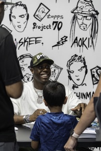 A man wearing a camo cap and white shirt is seated at a table, smiling as he interacts with a child in a blue shirt standing in front of him. The background features black-and-white illustrations and text, including words like 'Project 70' and 'AOK!'. Other people are partially visible, framing the interaction.