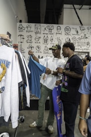Two people stand in a clothing store, examining T-shirts on racks. The wall behind them is decorated with various black and white illustrations, including faces, words, and numbers. One person is holding a blue shirt, while the other is holding a purple one. The atmosphere appears casual and relaxed.