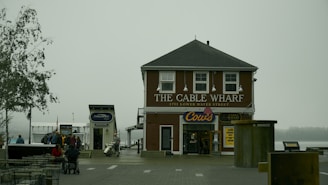 A foggy scene with a two-story building labeled 'The Cable Wharf' located at 1751 Lower Water Street. It has a brown exterior and white window frames. Below the main sign, there is another sign for 'Cows' indicating an ice cream shop. People are gathered around outside, with some sitting at tables and others walking nearby. A moored boat and a tree are visible to the left of the building.