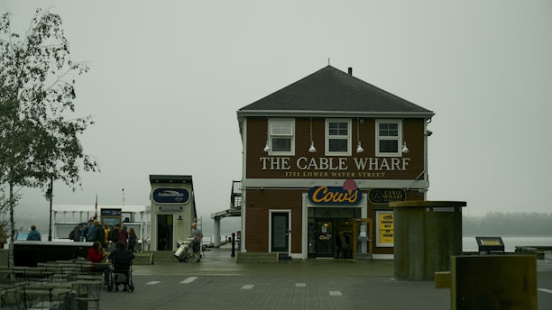 A foggy scene with a two-story building labeled 'The Cable Wharf' located at 1751 Lower Water Street. It has a brown exterior and white window frames. Below the main sign, there is another sign for 'Cows' indicating an ice cream shop. People are gathered around outside, with some sitting at tables and others walking nearby. A moored boat and a tree are visible to the left of the building.